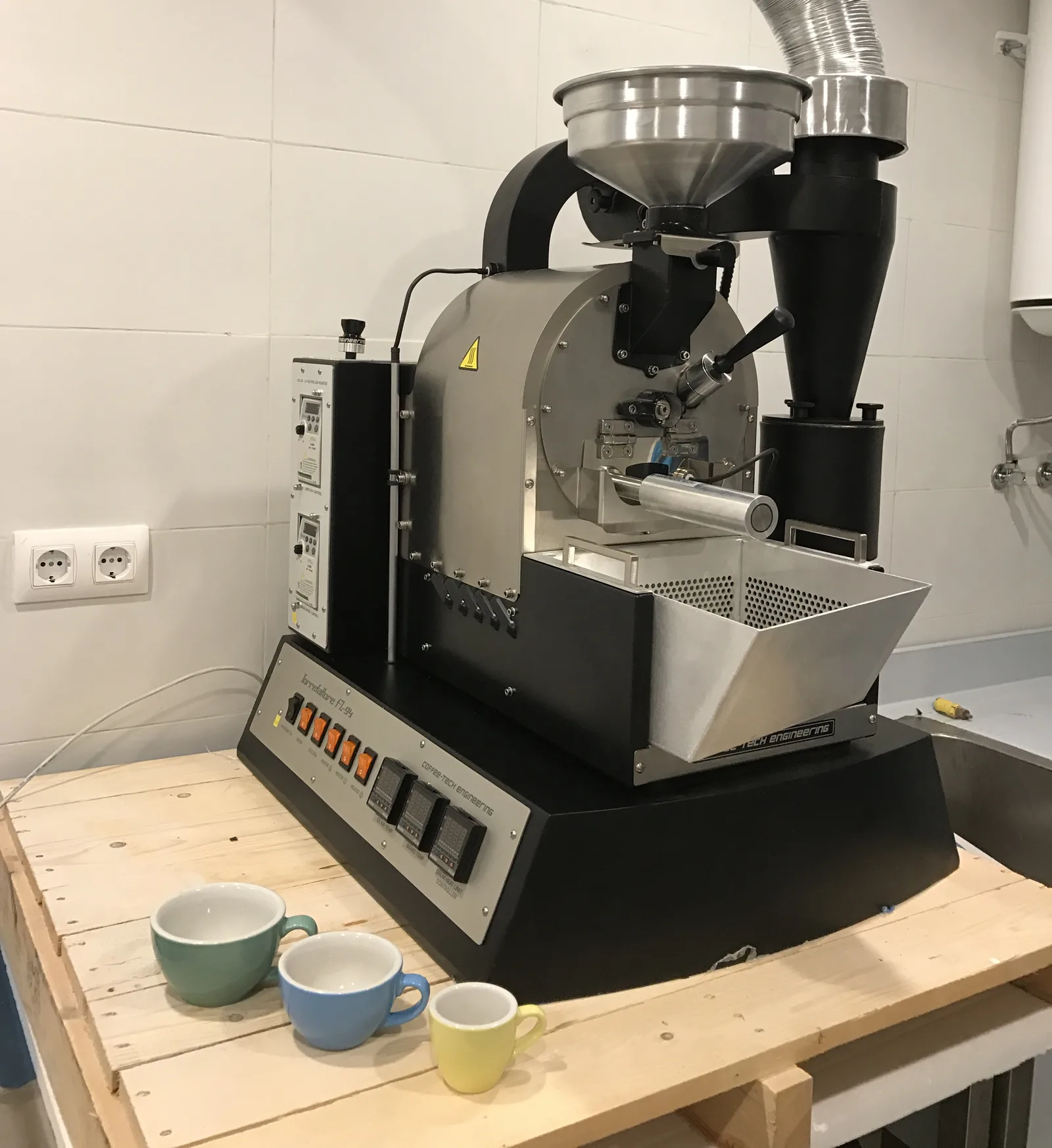 Tostadora de café Coffee-Tech FZ-94 recién instalada sobre una mesa de madera, con tres tazas de espresso delante, en la cocina alicatada del local de Boconó en Lavapiés, en diciembre de 2016