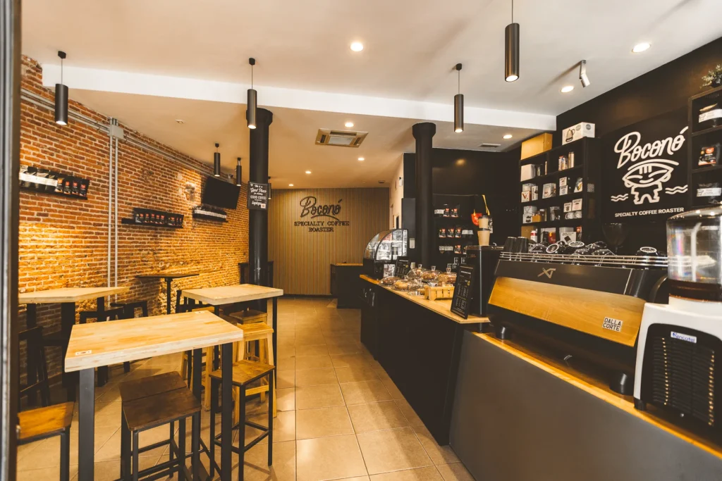 Interior de la cafetería de Boconó en Lavapiés, con la pared de ladrillo, las mesas altas de madera y la barra con el logotipo al fondo