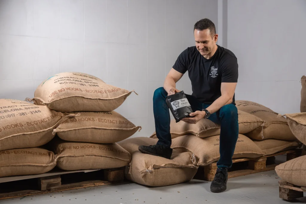 Carlos Núñez sostiene una bolsa de café de Boconó sentado sobre sacos de yute de café verde en el tostadero de Madrid