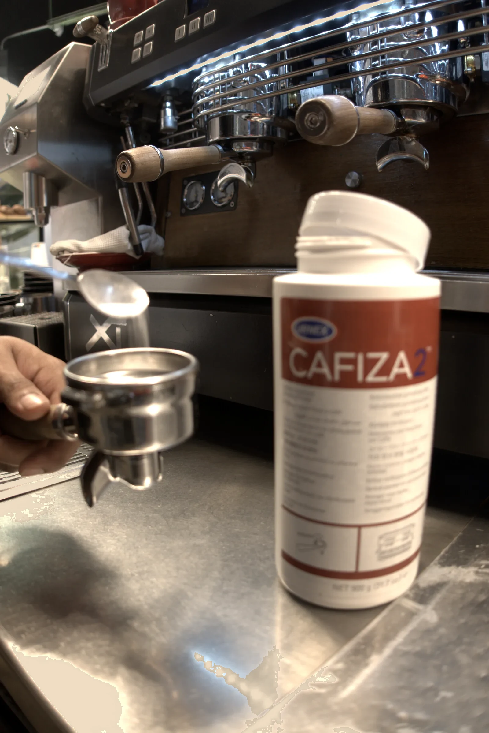 Un barista sostiene el portafiltros con la cesta ciega delante de la máquina de espresso del tostadero de Boconó, con el bote de Cafiza 2 de Urnex en la barra