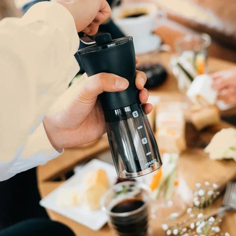 Una persona muele café con el molinillo manual sobre una mesa de desayuno, con tazas y comida alrededor.
