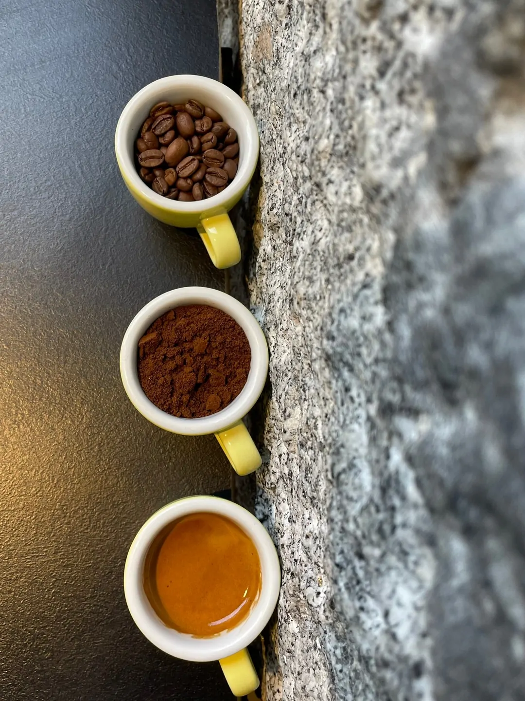 Tres tazas amarillas en fila sobre una barra negra junto a un pilar de granito: una con granos de café tostado, otra con café molido y otra con un espresso con crema