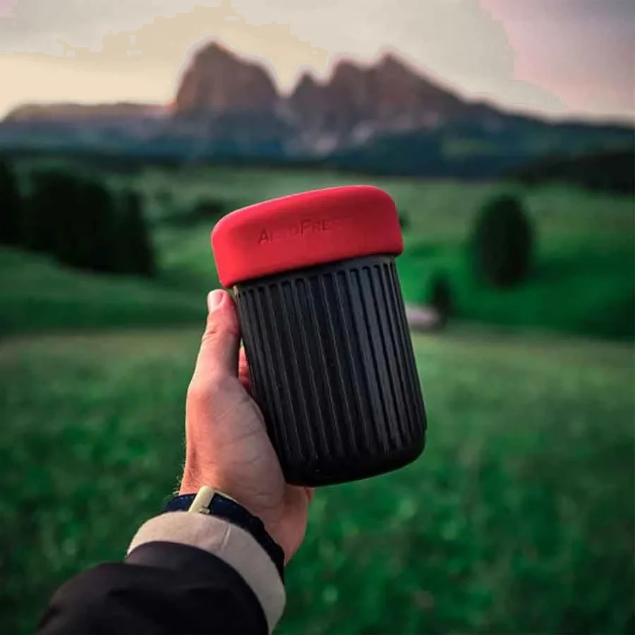 Una mano sujeta el vaso de viaje cerrado con su tapa roja, con el valle de fondo.