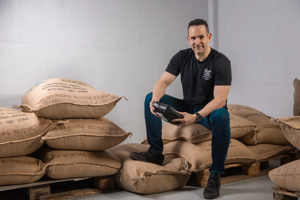 Carlos Núñez sentado sobre sacos de yute de café verde, con una bolsa de café de Boconó en las manos, en el tostadero de Madrid