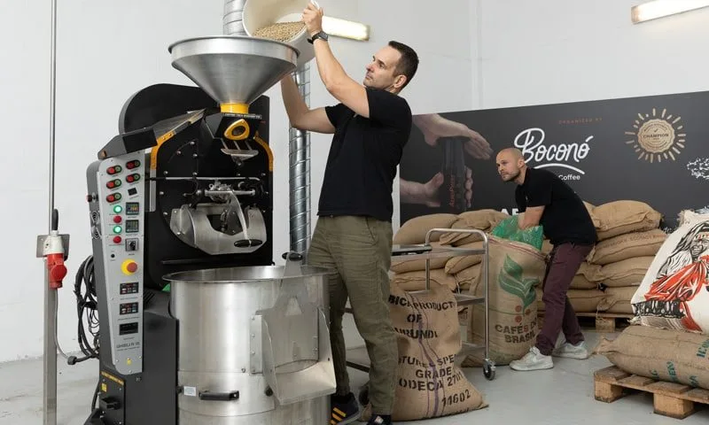 Carlos Núñez carga café verde en la tolva de la tostadora de Boconó, con Gastón Saldivia al fondo con un saco y los sacos de café verde de Burundi y de Brasil