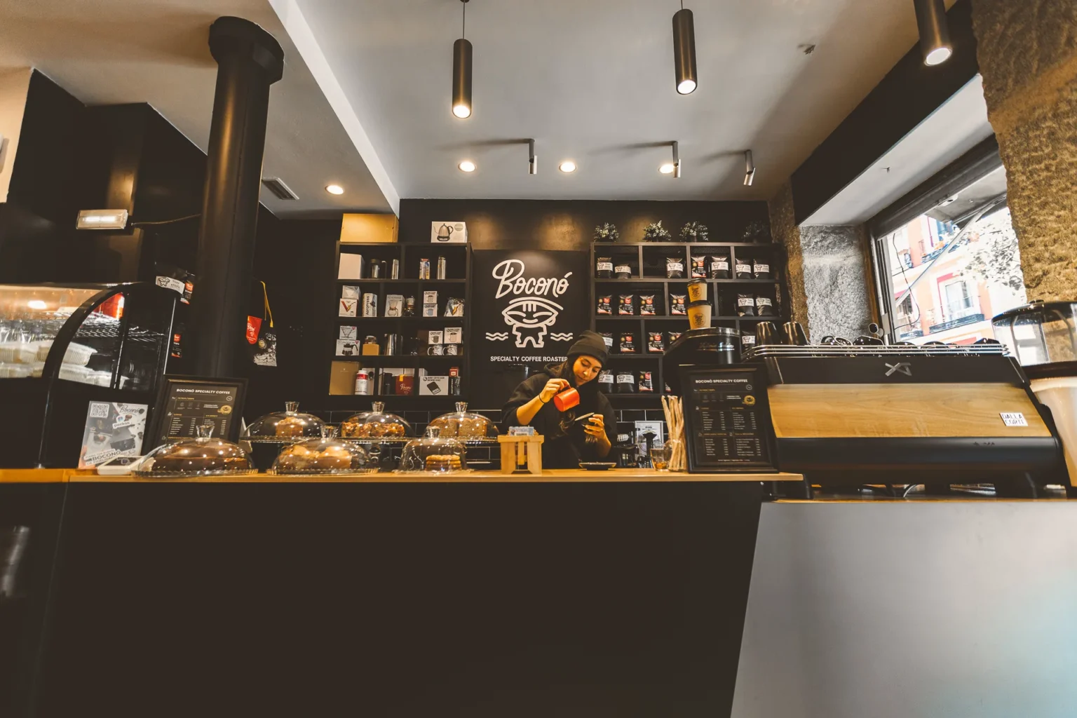 Una barista prepara un café con leche en la barra de Boconó en Lavapiés, con el logotipo de la marca pintado en la pared y las bolsas de café en las estanterías