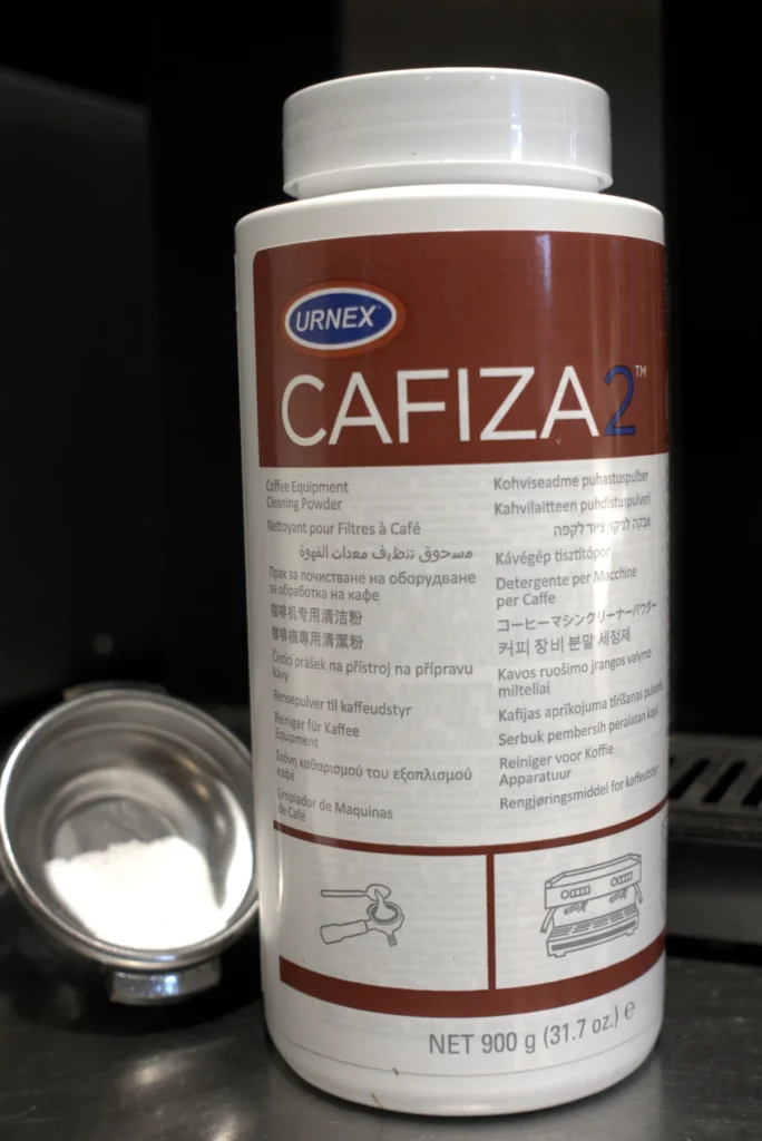Bote de 900 g de Cafiza 2 de Urnex junto a la cesta ciega del portafiltros con la dosis de detergente dentro