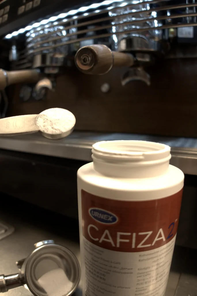 Cuchara medidora con la dosis de Cafiza 2 en polvo sobre el bote abierto, con el portafiltros y la cesta ciega al lado y la máquina de espresso al fondo