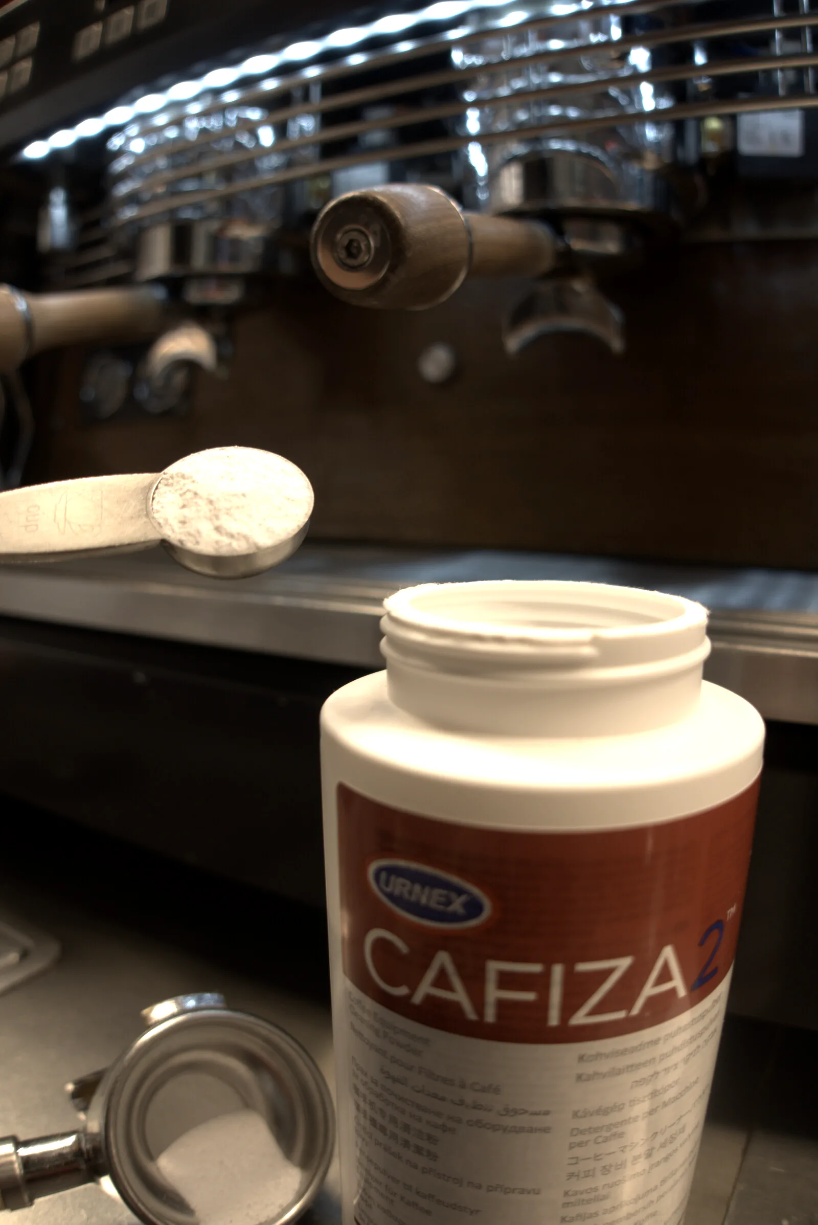 Cuchara medidora con la dosis de Cafiza 2 en polvo sobre el bote abierto, con el portafiltros y la cesta ciega al lado y la máquina de espresso al fondo
